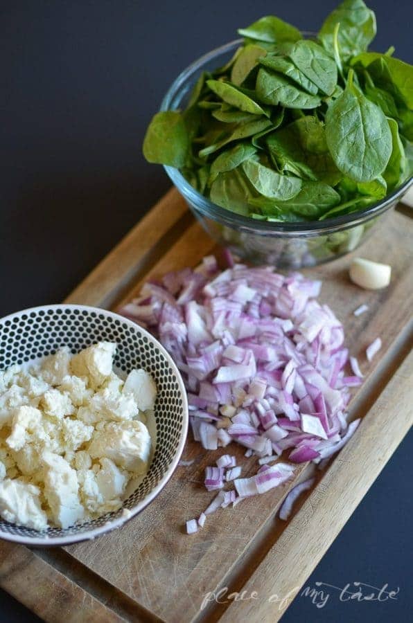 Fresh spinach in a glass bowl, crumbled feta cheese and diced red onion on a wooden cutting board
