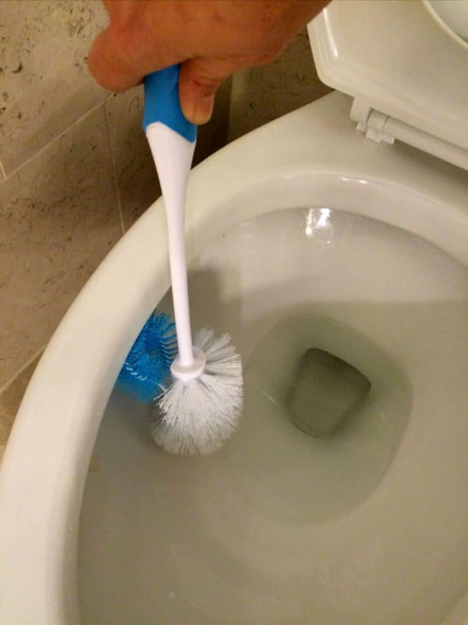 Gloved hand scrubbing the inside of a toilet bowl with a white toilet brush that has a blue under-rim brush
