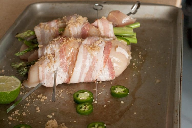 Bacon-wrapped stuffed chicken breasts on a metal baking tray with asparagus peeking out, sliced jalapeños and half a lime
