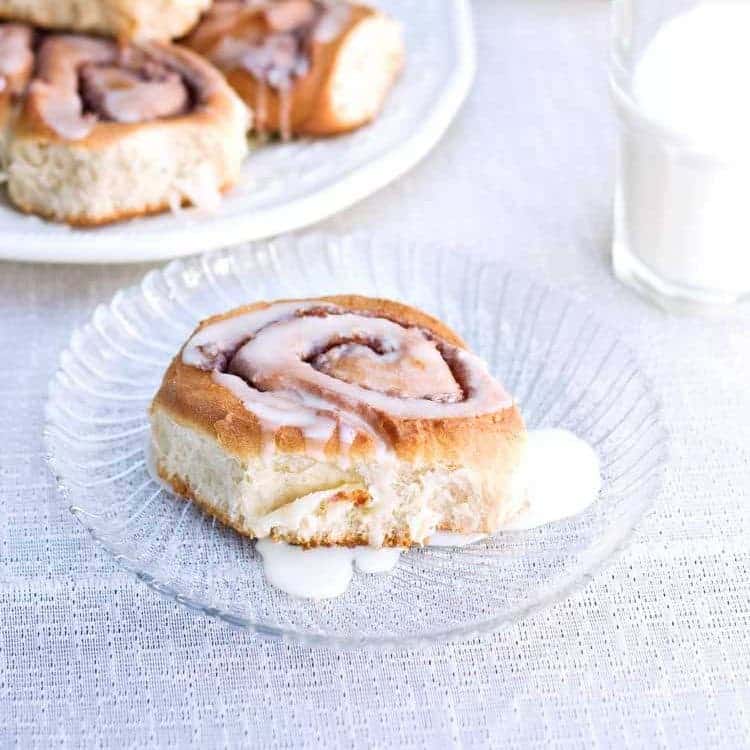 cinnamon roll on clear plate with white plate and stack of cinnamon rolls off to the side