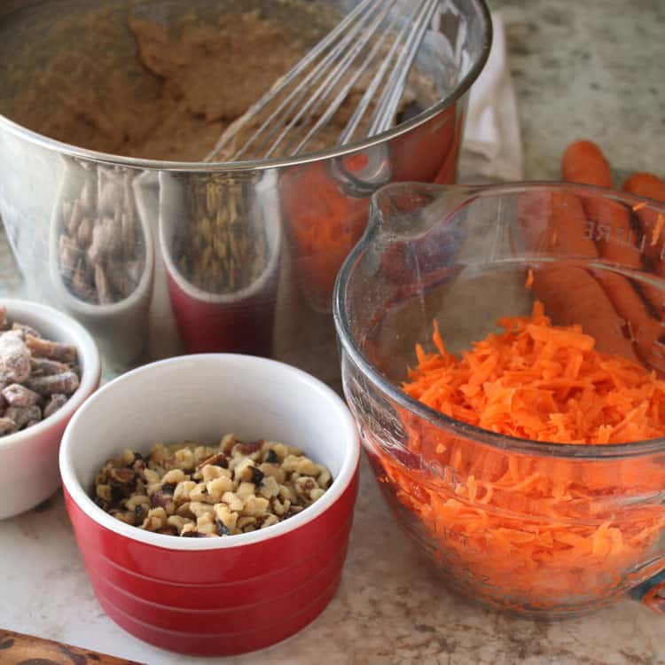 small red bowl containing nuts for best carrot cake recipe next to clear glass bowl of shredded carrots in front of stainless bowl