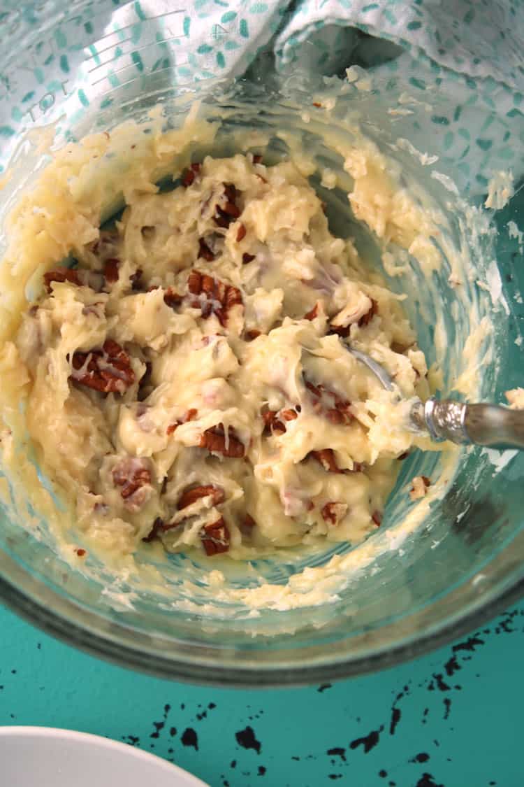 glass mixing bowl containing melted white chocolate, coconut, and pecans mixed together for coconut clusters with a spoon dipping in