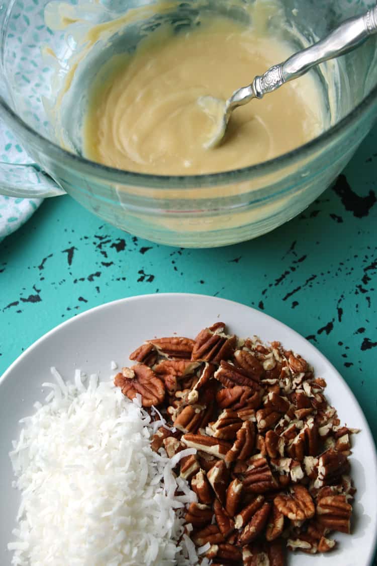 melted white chocolate in background for coconut clusters with plate of pecans and coconut flakes in foreground