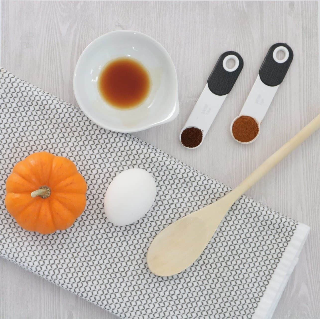 Ingredients for pumpkin patties including a mini pumpkin, an egg, vanilla extract, cinnamon, and cloves with a wooden spoon