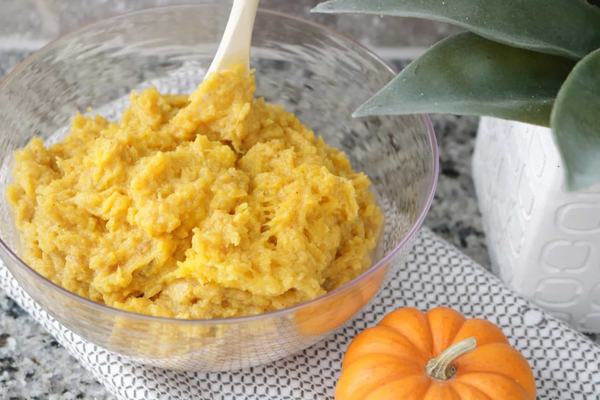Mashed pumpkin patty batter in a clear mixing bowl with a mini pumpkin on the side