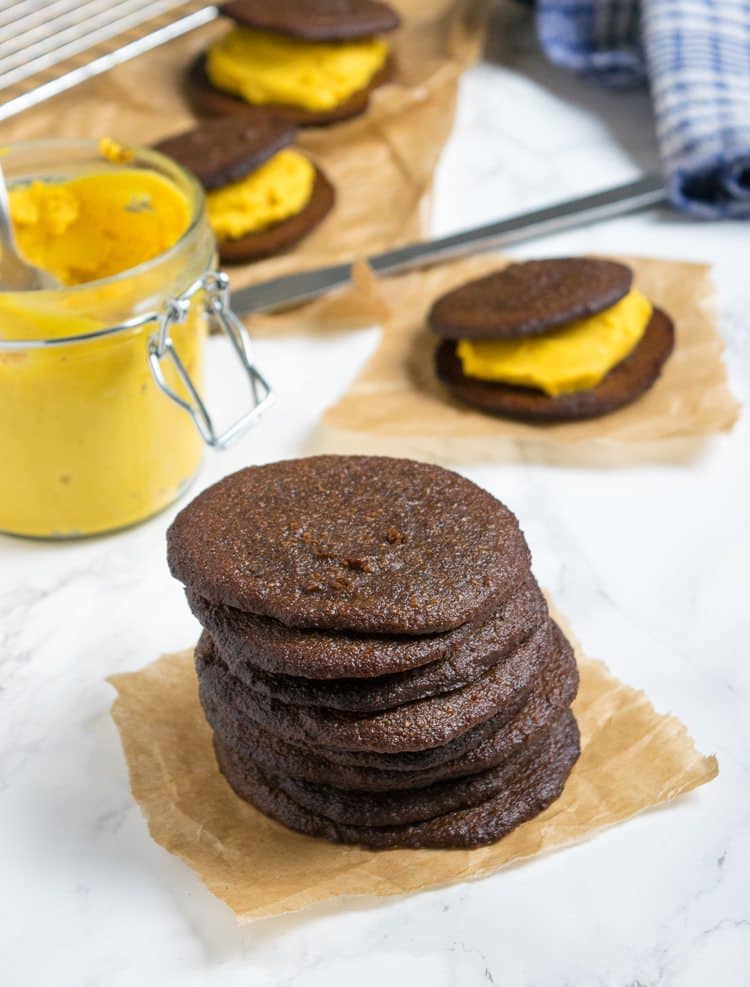 Stack of pumpkin whoopie pie cookies on parchment paper with a jar of pumpkin cream and filled whoopie pies behind