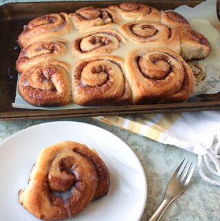 homemade cinnamon rolls sitting on a baking sheet in the background with a single cinnamon roll on a white plate in the foreground