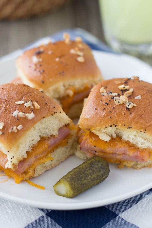 Ham and cheddar potluck sandwiches on sweet rolls topped with poppy seeds and minced onion, served on a white plate with a pickle