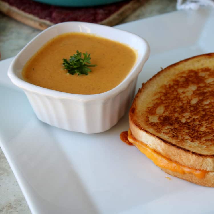 carrot soup with parsley garnish in white bowl sitting on a white plate next to a grilled cheese sandwich