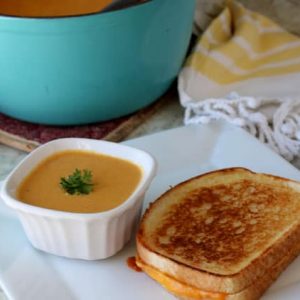 soup in white dish next to sandwich on white plate with carrots, a blue pot and yellow and white towel in the background