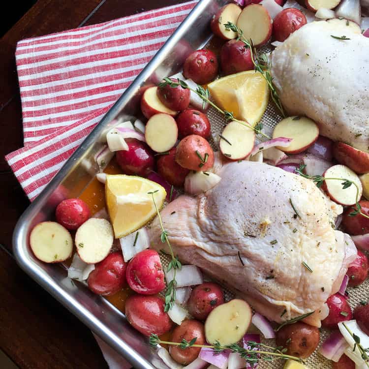 sheet pan chicken with raw herb-roasted chicken, red potatoes, lemon, kale and sprigs of thyme on a sheet pan on top of a striped towel