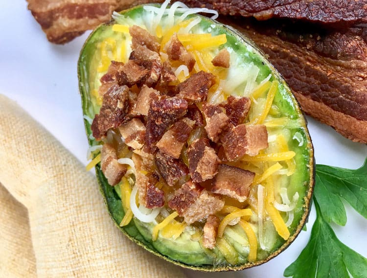 half of a baked avocado containing melted cheese and bacon bits next to strips of bacon 