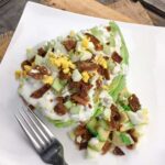 Permalink to: Keto Salad - Wedge Salad with Homemade Blue Cheese Dressing keto salad on a white plate consisting of iceberg lettuce, bacon, and eggs next to a fork