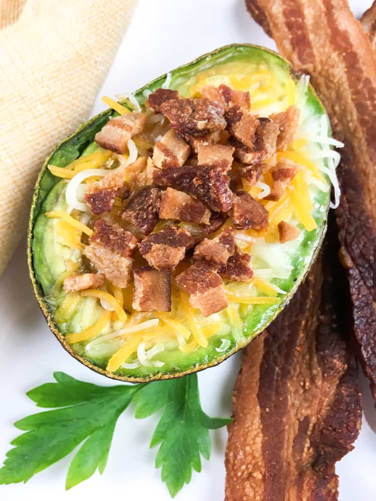 overhead view of avocado half stuffed with melted cheese and bacon bits next to strips of bacon