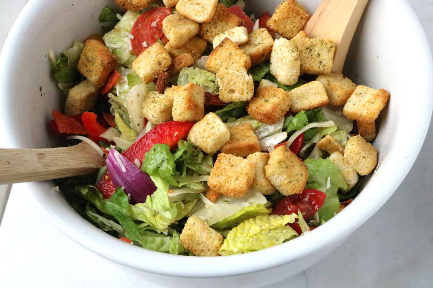close-up of Italian salad topped with croutons in a white bowl with salad forks. 
