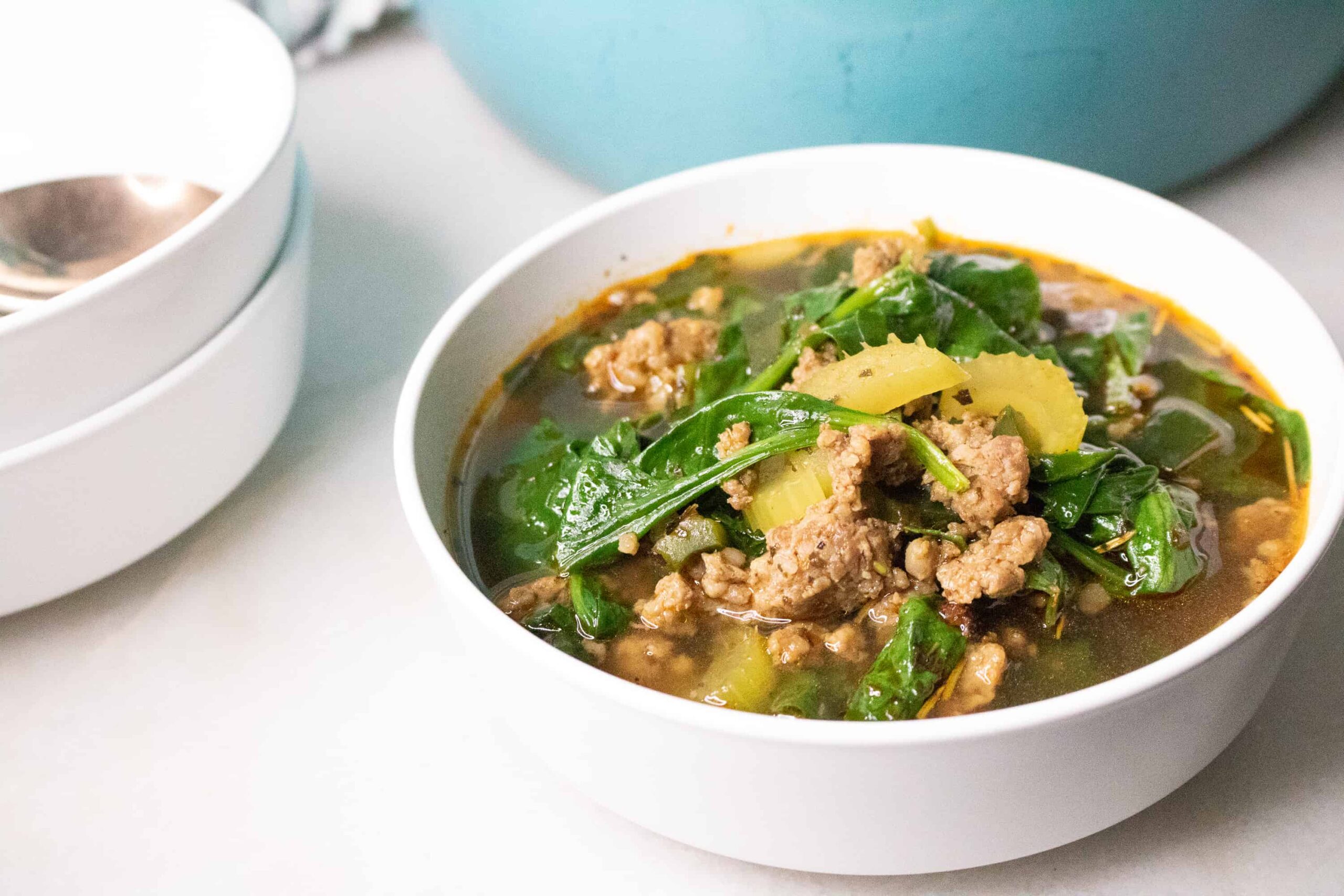 Bowl of keto sausage soup with spinach and celery beside a teal Dutch oven
