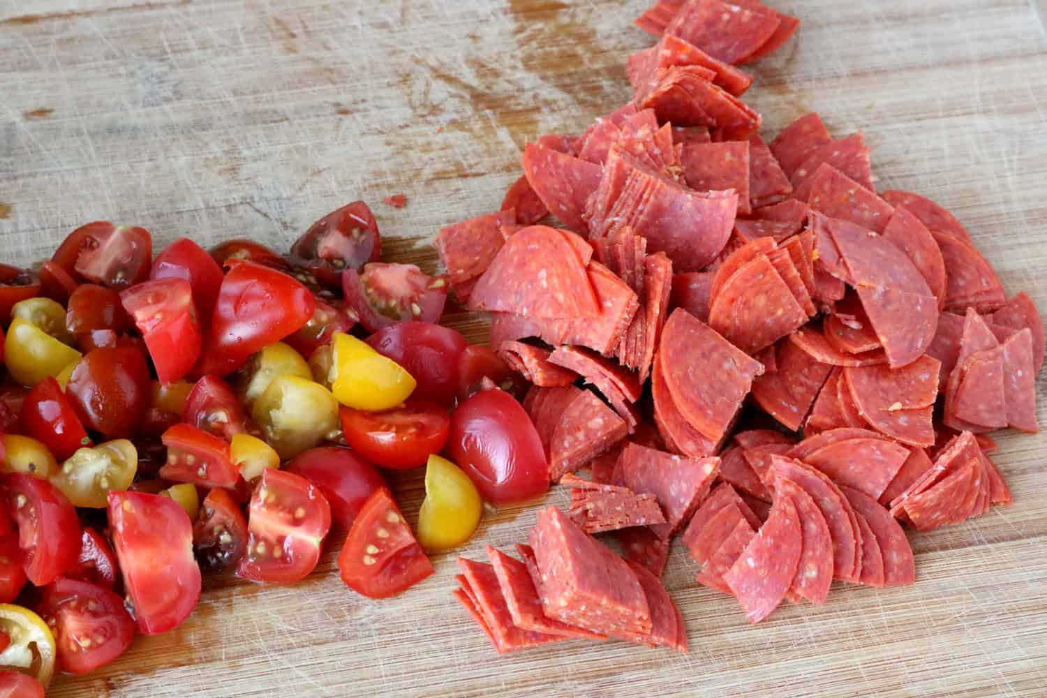 Quartered cherry tomatoes and pepperoni shown on a wooden cutting board.