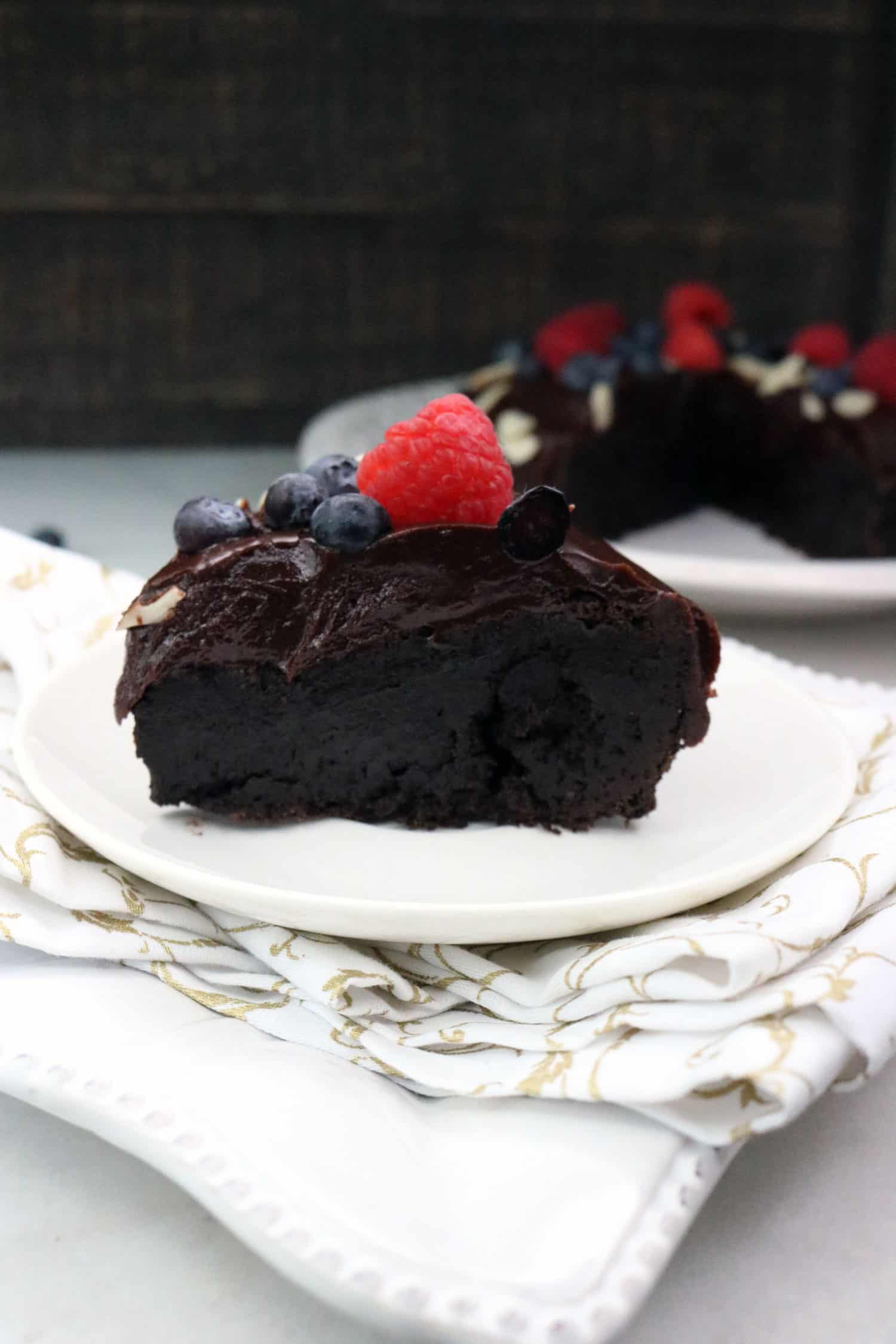 Slice of Gluten Free Cake - Flourless Chocolate Cake with Dark Chocolate Ganache topped with a raspberry and blueberries on a white plate