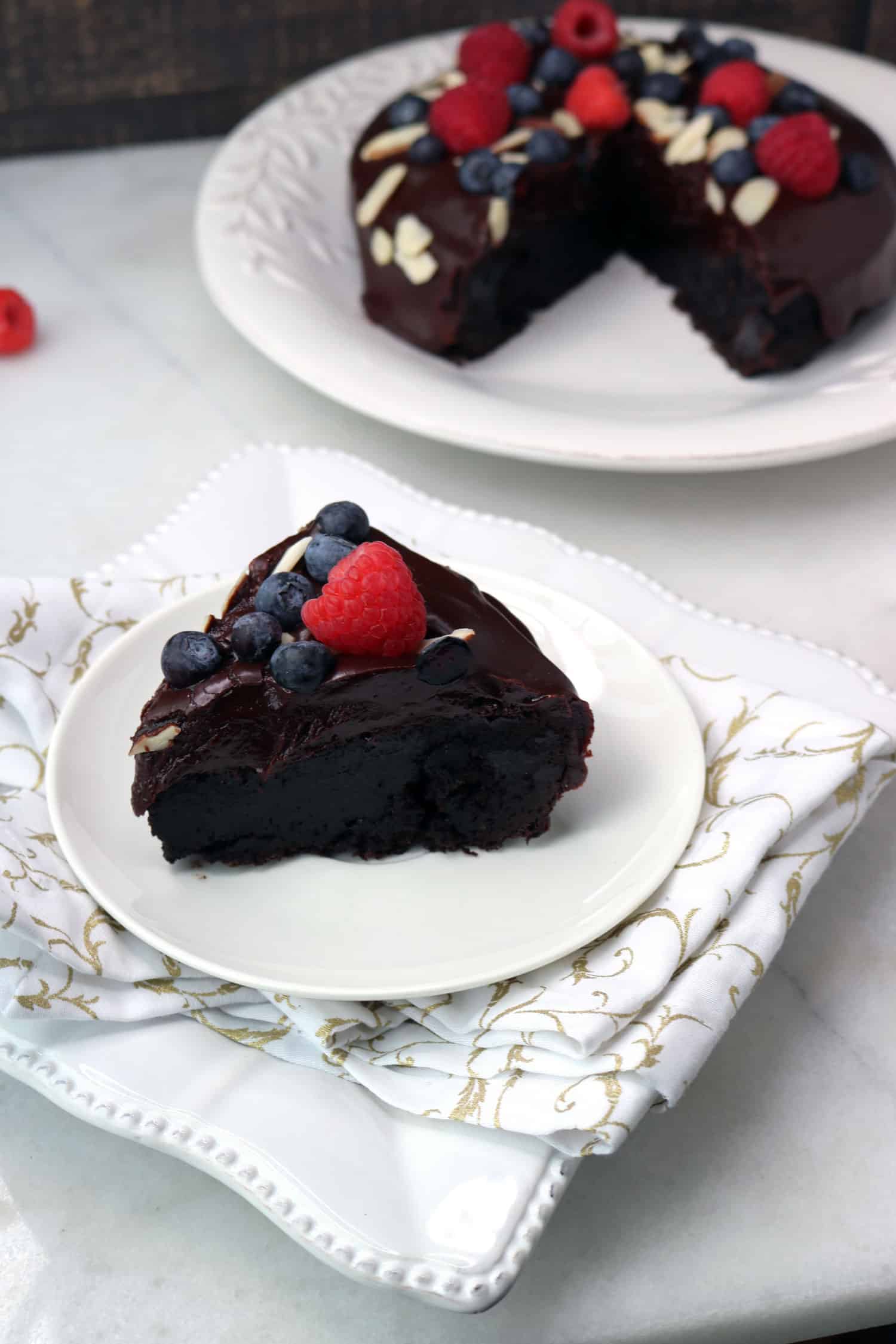 Gluten Free Cake - Flourless Chocolate Cake with Dark Chocolate Ganache topped with raspberries and blueberries sitting on a white plate with a slice cut out sitting on a white plate infront