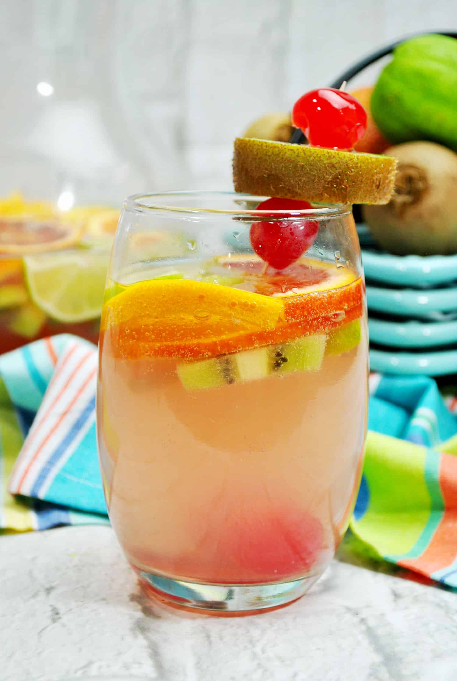 up close picture of clear glass with peach color tropical fruity sangria recipe in it garnished with oranges, cherries and kiwi
