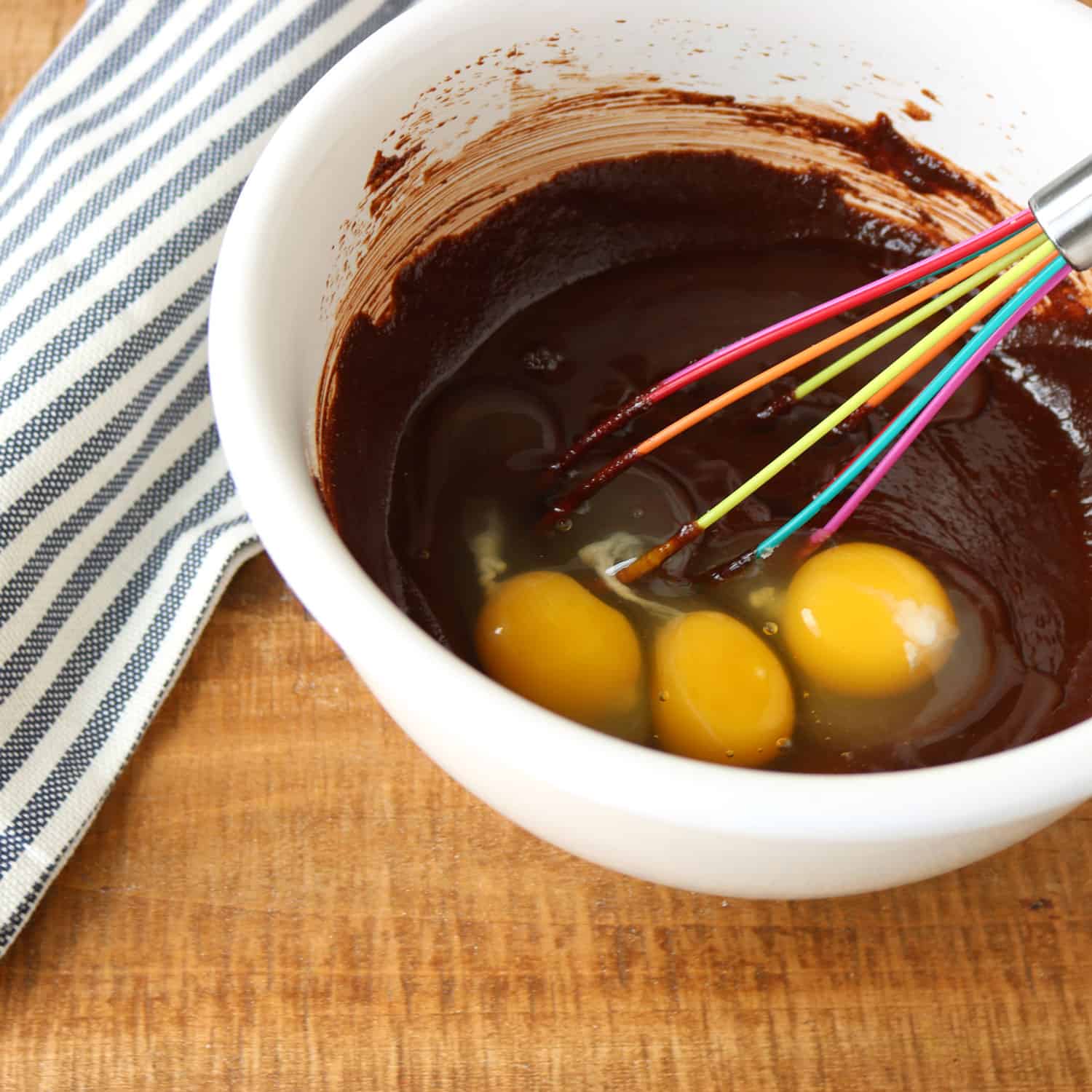 chocolate gluten free cake mixture with three raw eggs in a white bowl with a rainbow-colored whisk