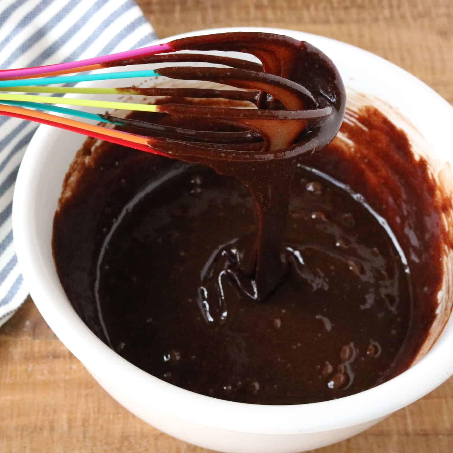 chocolate gluten free cake mixture dripping from a rainbow-colored whisk into a bowl of melted chocolate below
