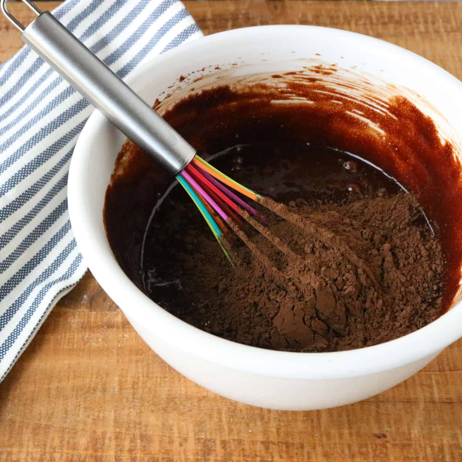 chocolate gluten free cake mixture with raw cocoa powder on top in a white bowl with a rainbow-colored whisk