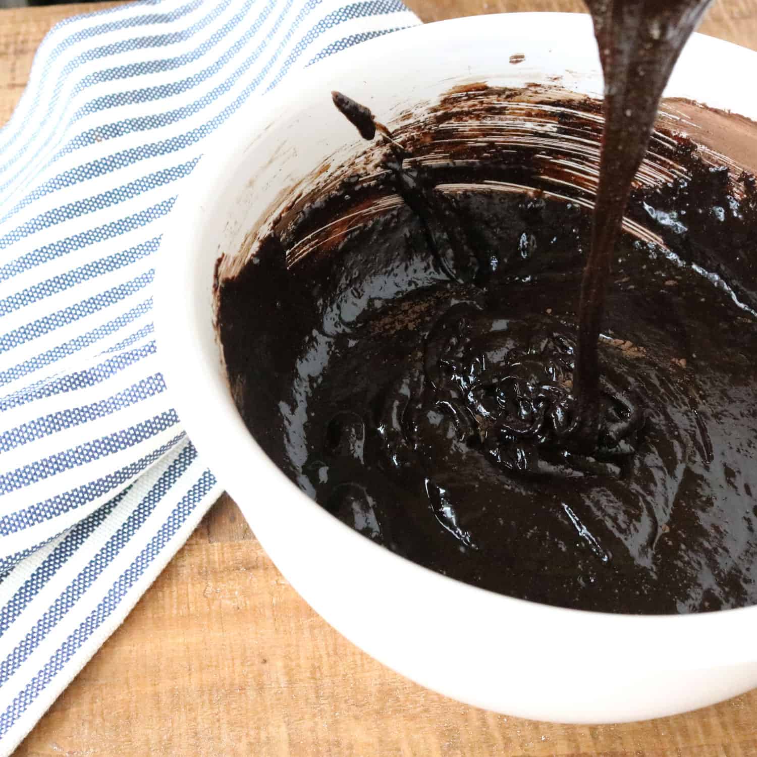 chocolate gluten free cake mixture pouring into a white bowl next to a blue and white striped kitchen towel