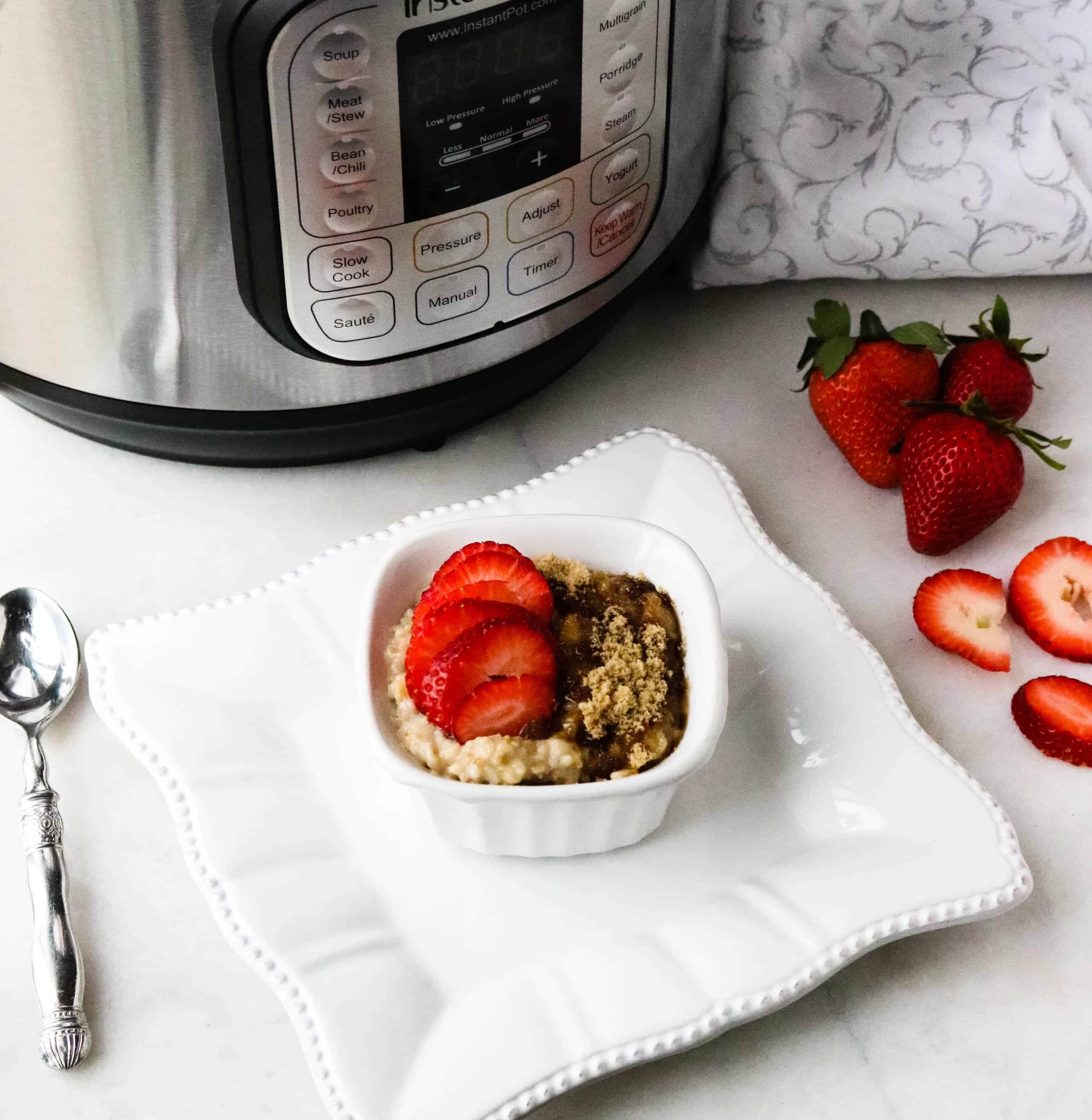 Instant Pot steel cut oats topped with sliced strawberries and brown sugar and served in a white bowl placed on a square white plate with a silver spoon and three whole strawberries on the table