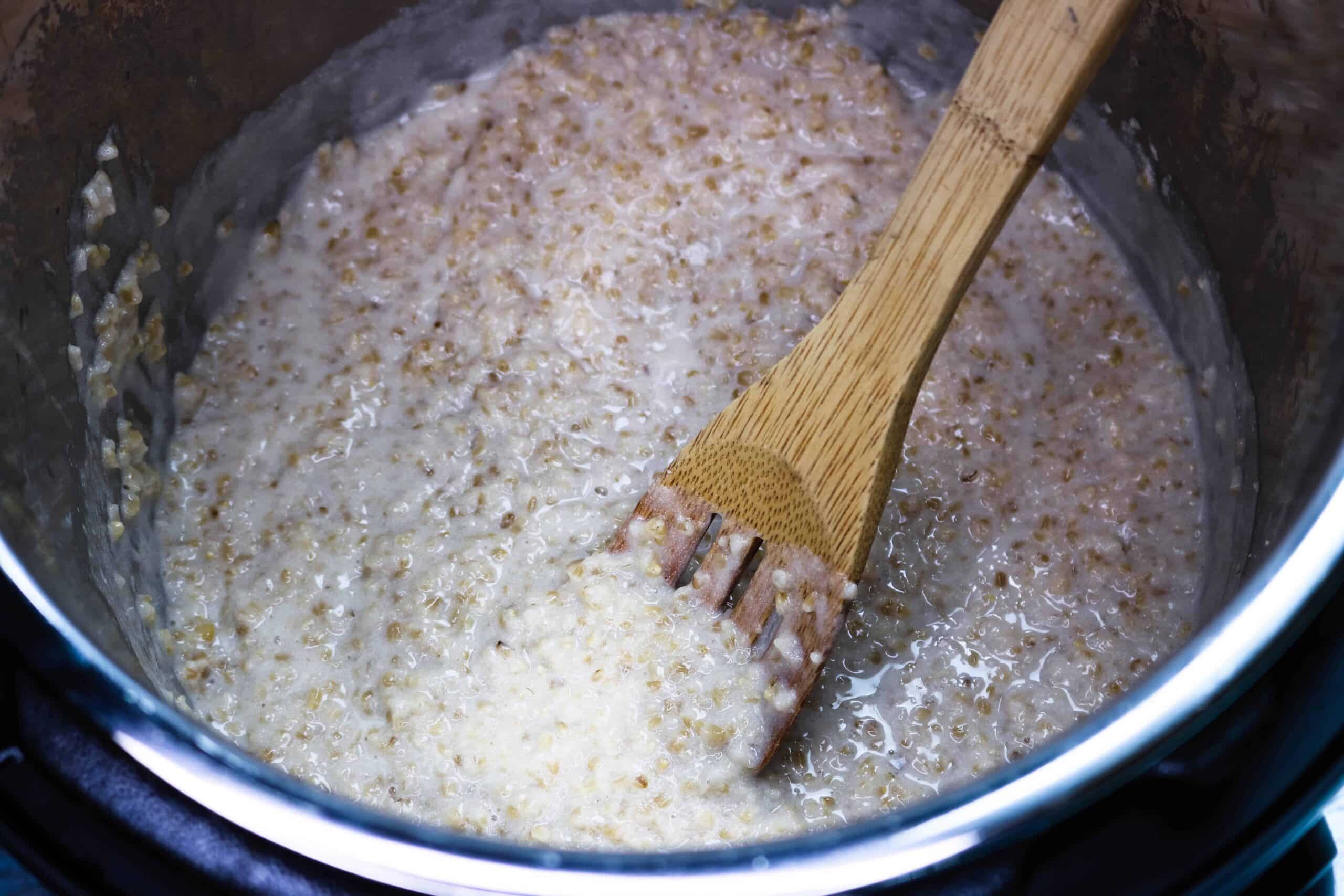 cooked steel cut oats inside the Instant Pot being stirred with a wooden spoon