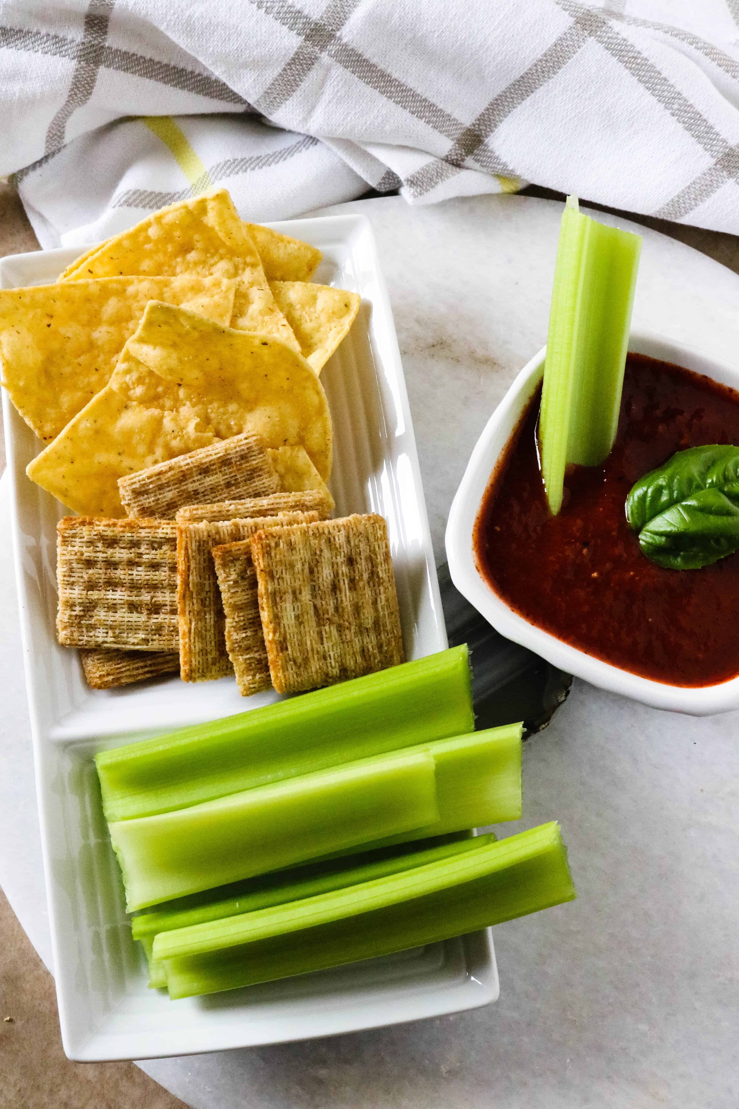White plate with crackers, tortilla chips and celery in foreground. Small white bowl of Roasted Red Pepper dip to the right