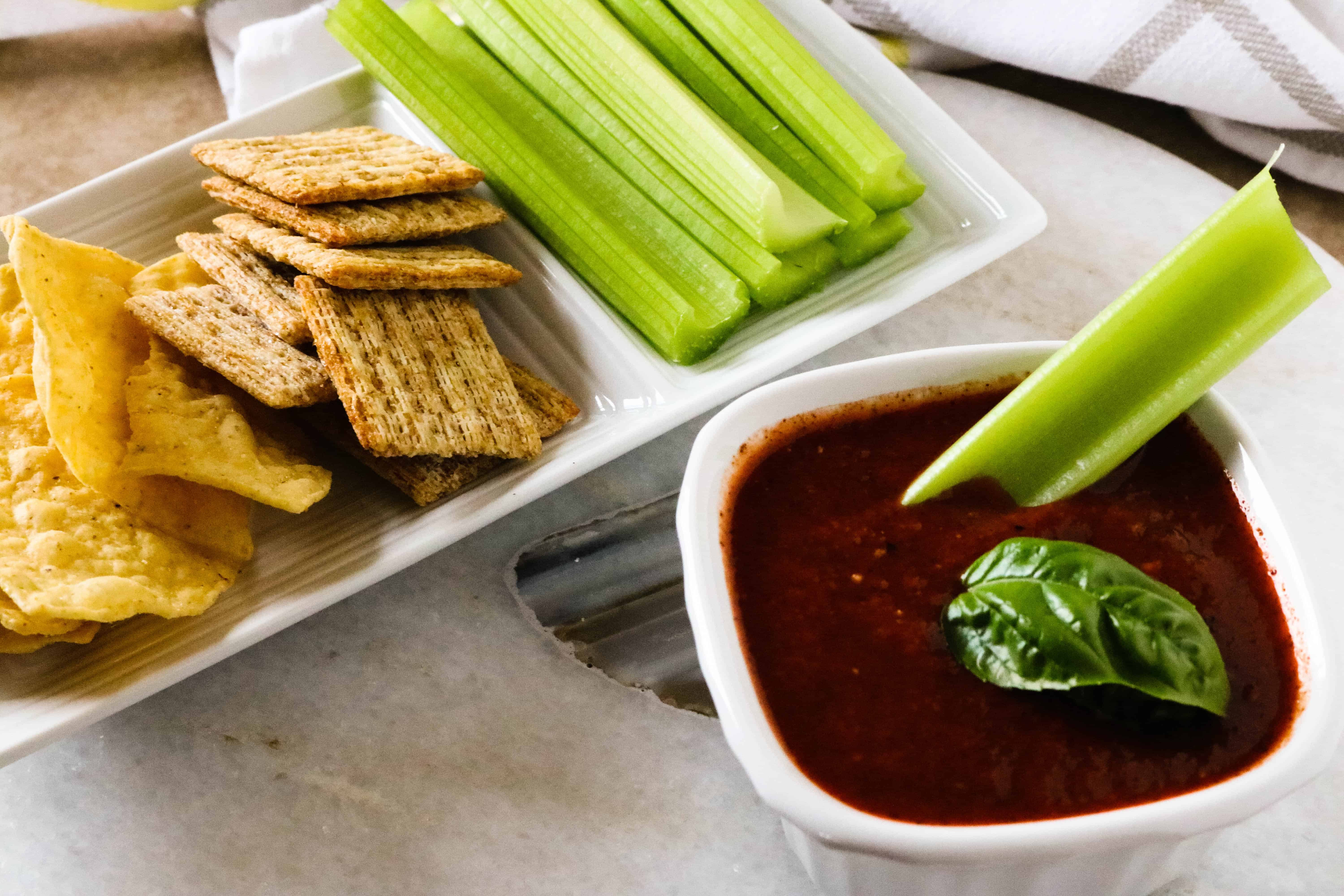 rectangular white plate with celery, triscuit crackers and tortillas in background and white ceramic bowl of roasted red pepper dip in foreground