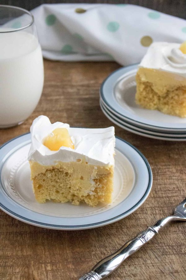 A slice of pineapple poke cake topped with whipped topping and a pineapple chunk, with more slices and a glass of milk behind it