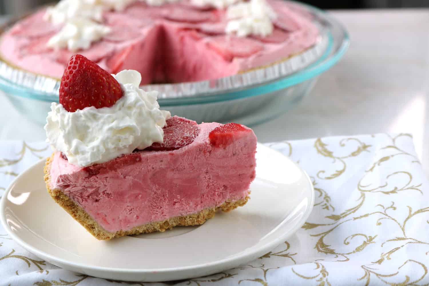 a slice of boozy frozen strawberry daiquiri pie served on a white plate and topped with whipped cream and a strawberry