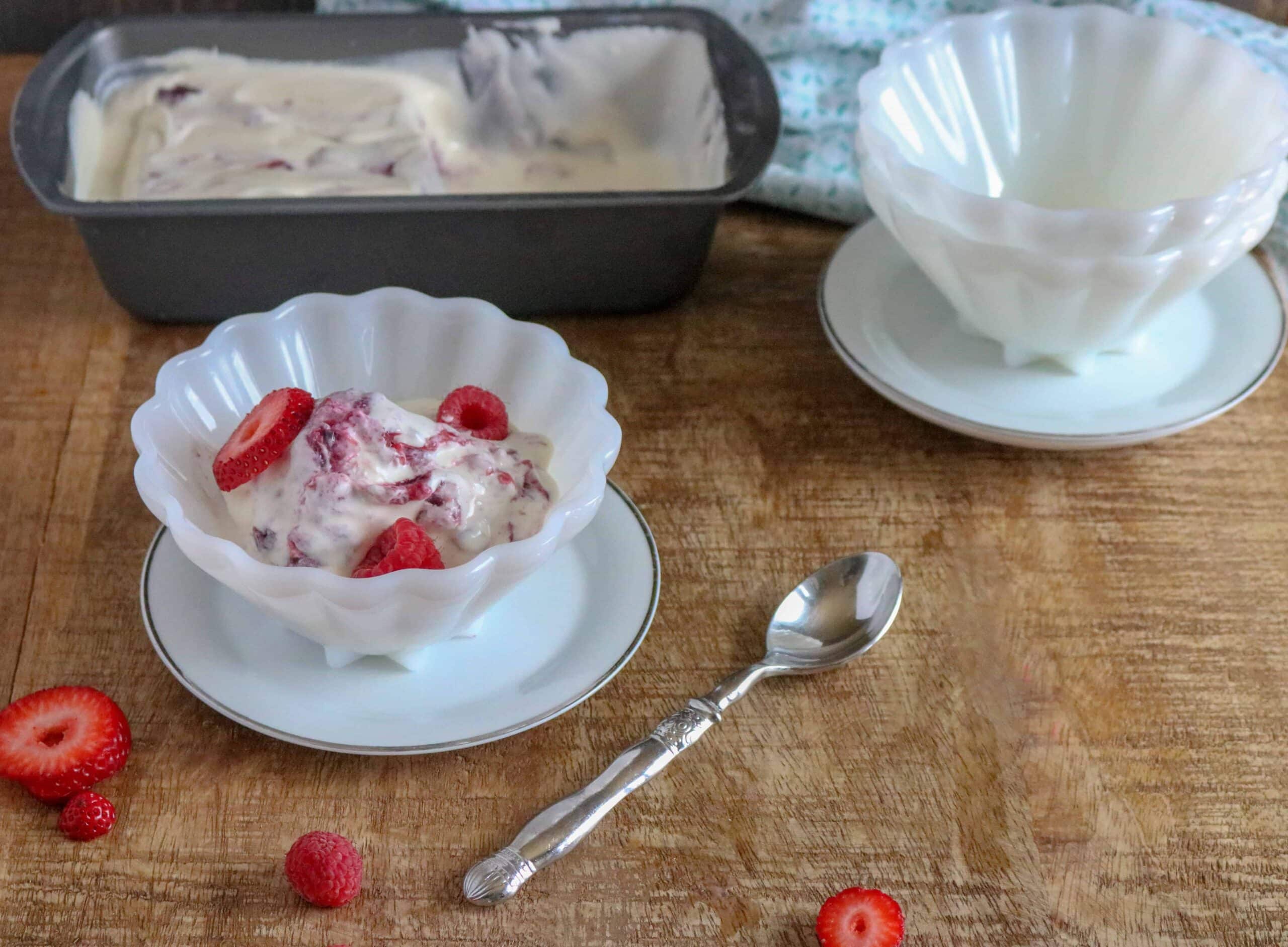 Berry Pie No-Churn Ice Cream in a white scalloped bowl and a white saucer sitting on a wooden table next to a spoon, berry slices, a pan of Berry Pie No-Churn Ice Cream and scalloped white bowls