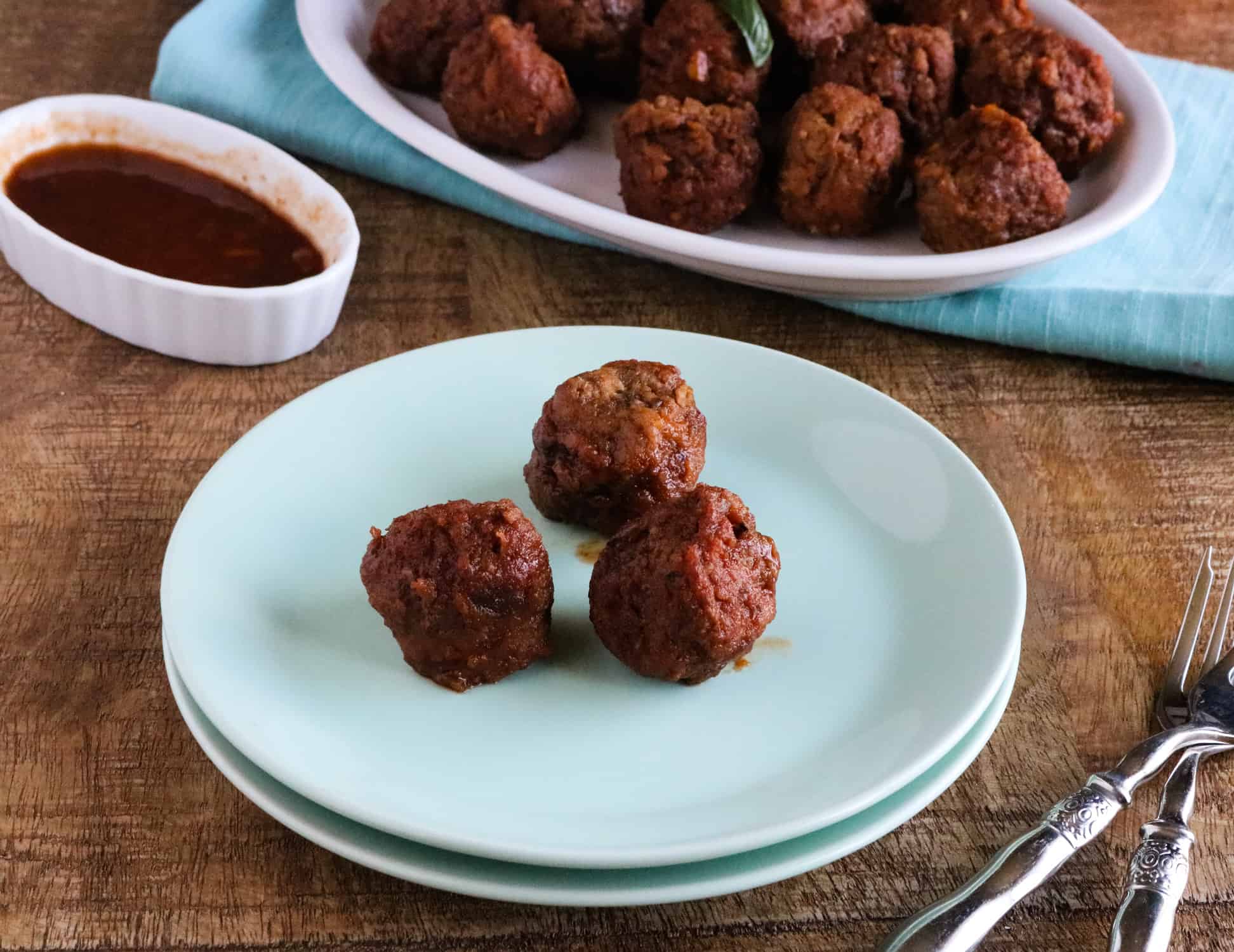Three Instant Pot Orange BBQ Meatballs served on a blue plate next to a white platter holding the rest of the meatballs, with a ceramic dish of orange BBQ sauce on the side