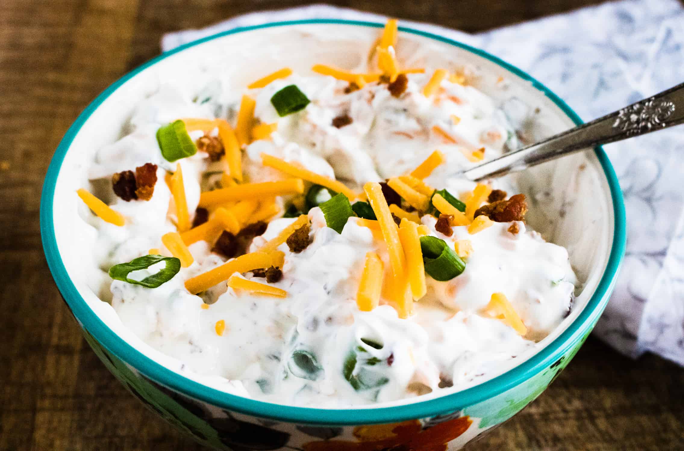 close up view of loaded baked potato dip in a floral serving bowl being mixed with a silver spoon