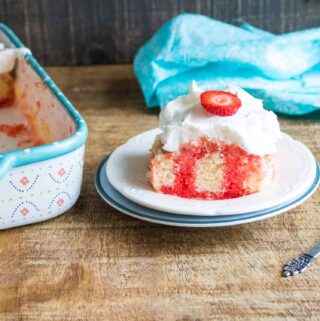 Completed Strawberry Poke Cake in a blue print sheet pan sitting next to a slice served on a white dessert plate with a blue rim