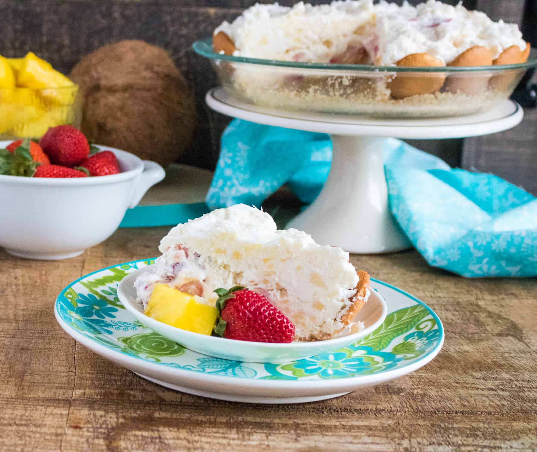 piece of no bake tropical cream pie on a small with dish sitting on a blue and green liner plate with white bowl of strawberries in background next to a white cake stand holding the pie plate of no bake tropical cream pie