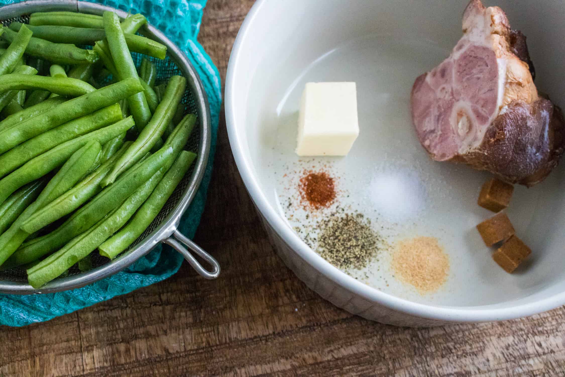 top-down view of fresh green beans in a colander next to a white ceramic bowl containing ham hock, butter, bouillon cubes, garlic powder, cayenne, salt and pepper 