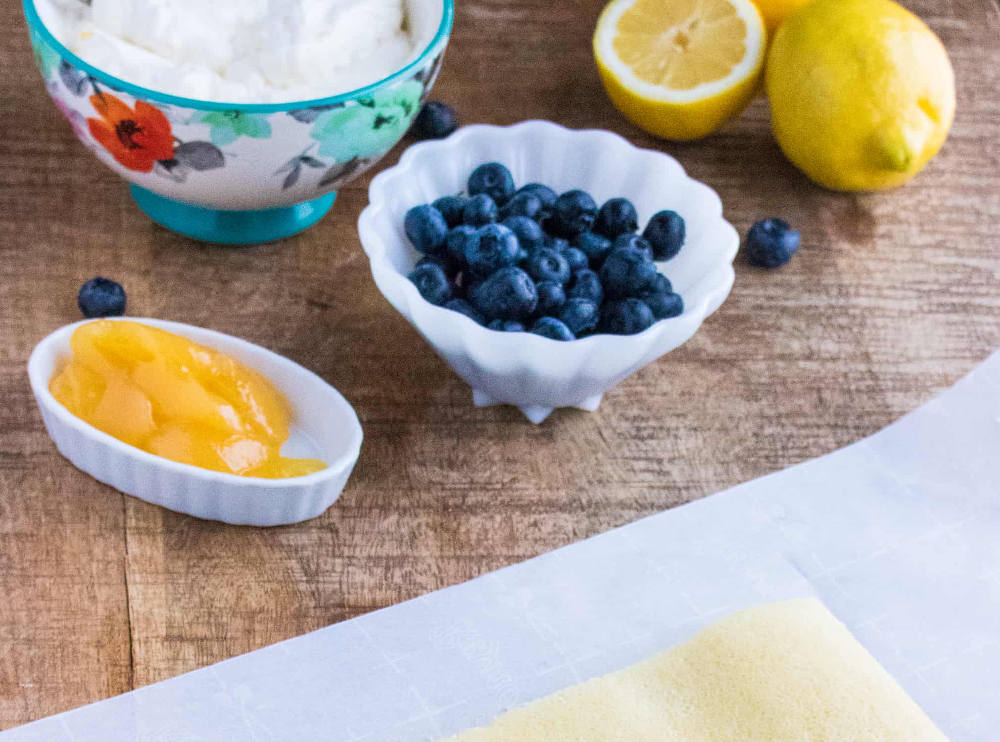 lemon curd in a white bowl, blueberries in a white scalloped bowl, whipped cream in a floral bowl on a wooden table next to lemons and a piece of parchment paper