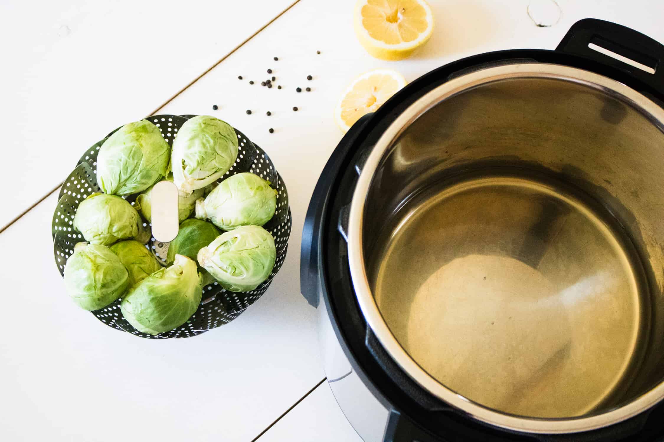 top-down view of instant pot with chicken broth inside, sitting next to steamer basket filled with Brussels sprouts, with whole peppercorns and a lemon cut in half placed behind them on the counter