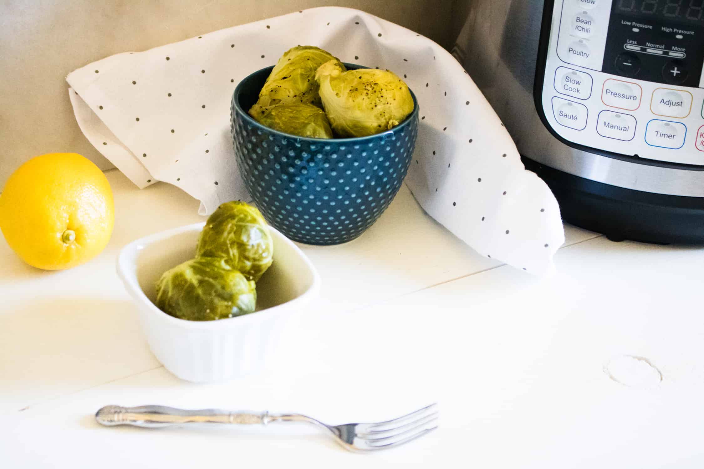 two bowls of instant pot lemon pepper brussels sprouts next to a whole lemon, polka dot cloth napkin, and silver fork
