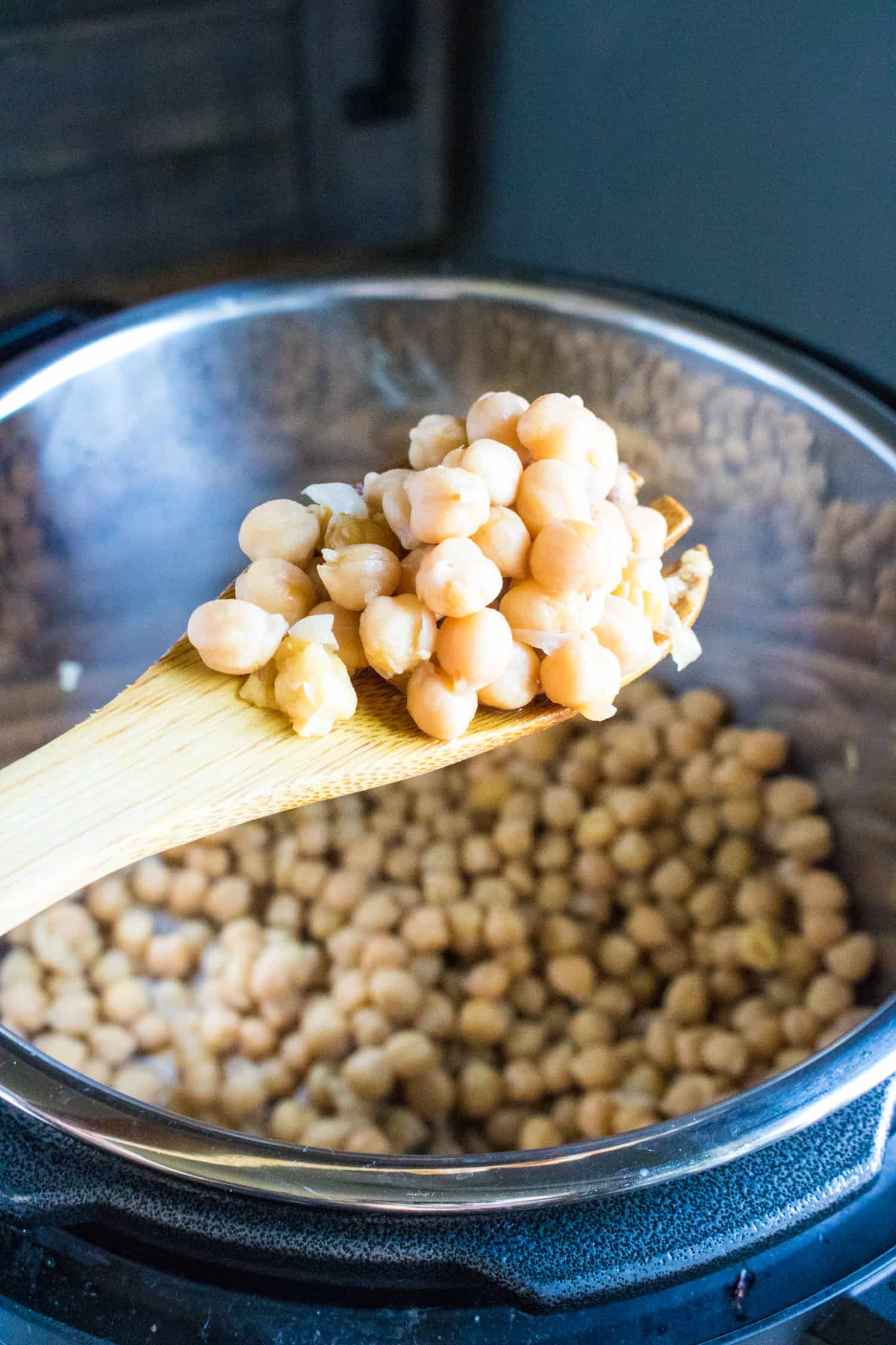wooden spoon showing cooked chick peas for Roasted Red Pepper Hummus with instant pot in background full of chick peas