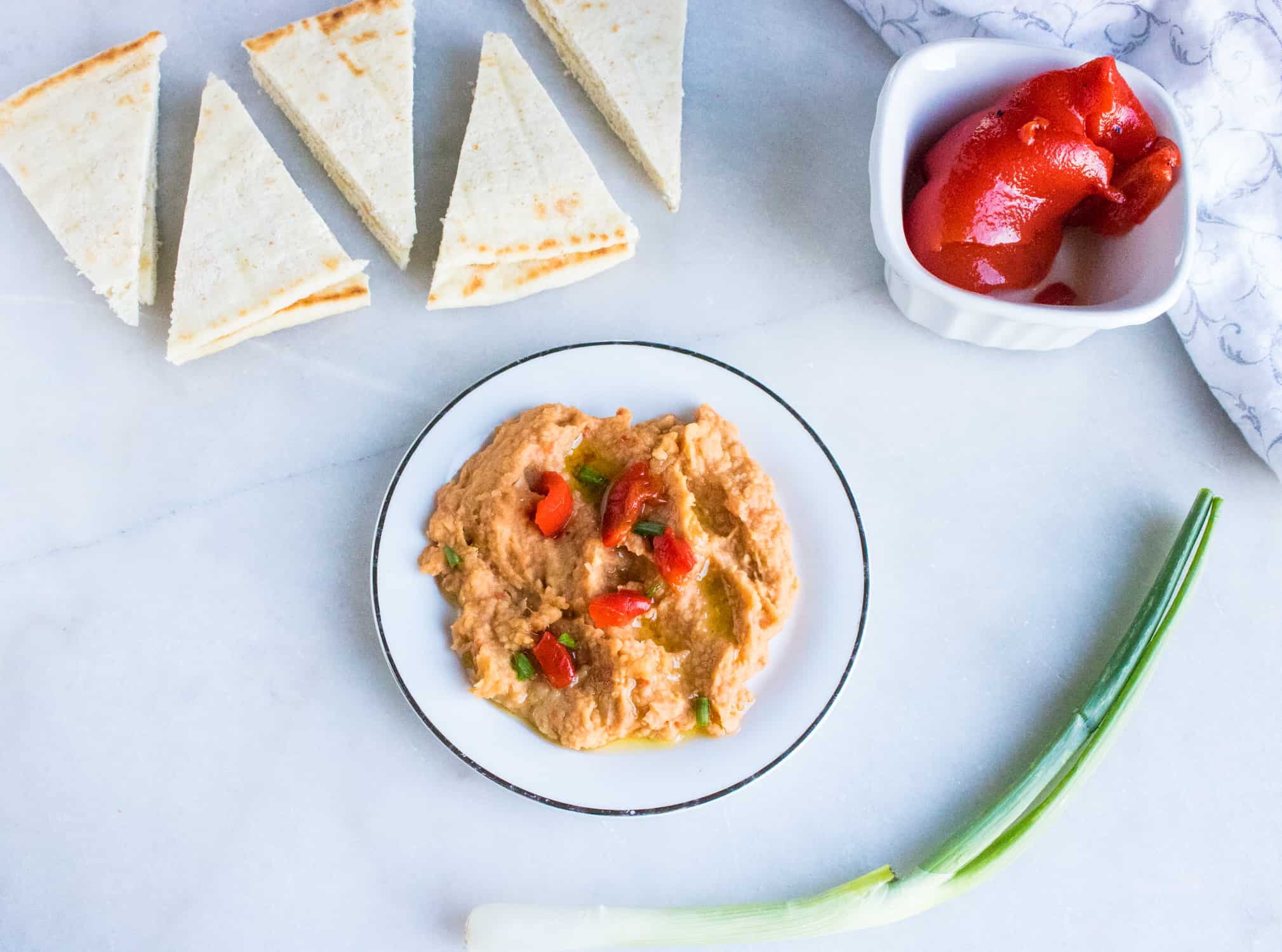 sliced triangles of pita bread in background with white bowl in foreground with Roasted Red Pepper Hummus in it with white ceramic dish with whole roasted red pepper in it and green onion laying in front