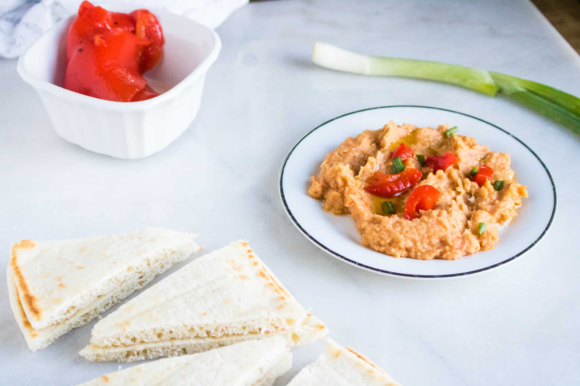 white ceramic dish in background with whole roasted red pepper in it. sliced triangles of pita bread in foreground sitting next to white bowl with Roasted Red Pepper Hummus in it with green onion laying behind