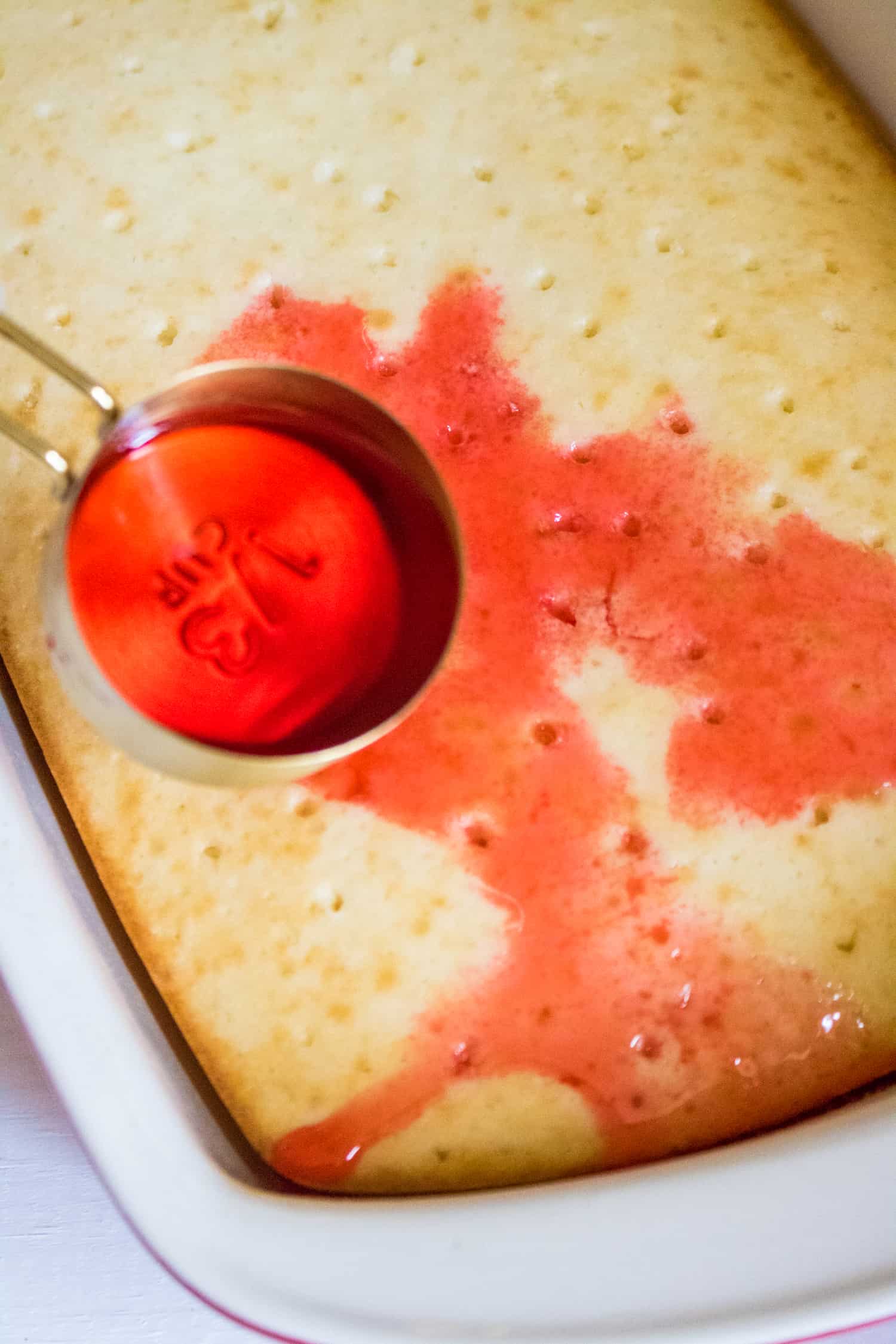 Cherry syrup being poured over baked white cake and filling the small poked holes 