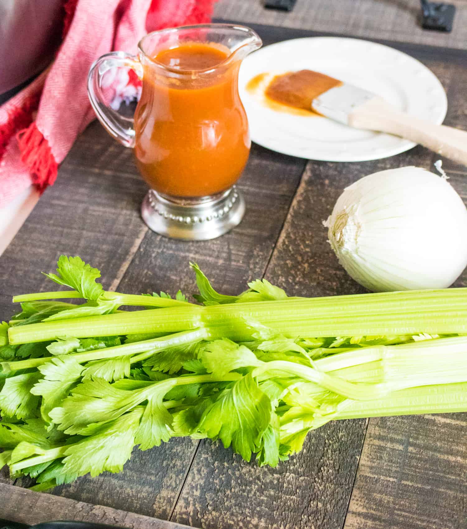barbecue Sauce in a clear glass pitcher next to celery stalks, an onion, and a BBQ brush on a wooden serving tray