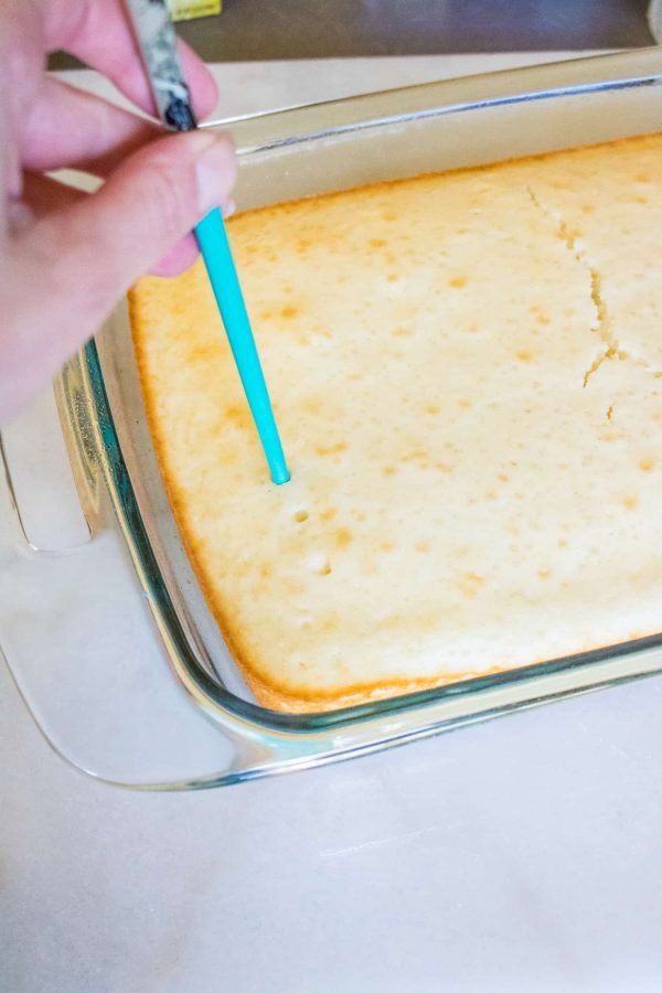 Poking holes in a baked white cake in a glass pan with a blue skewer