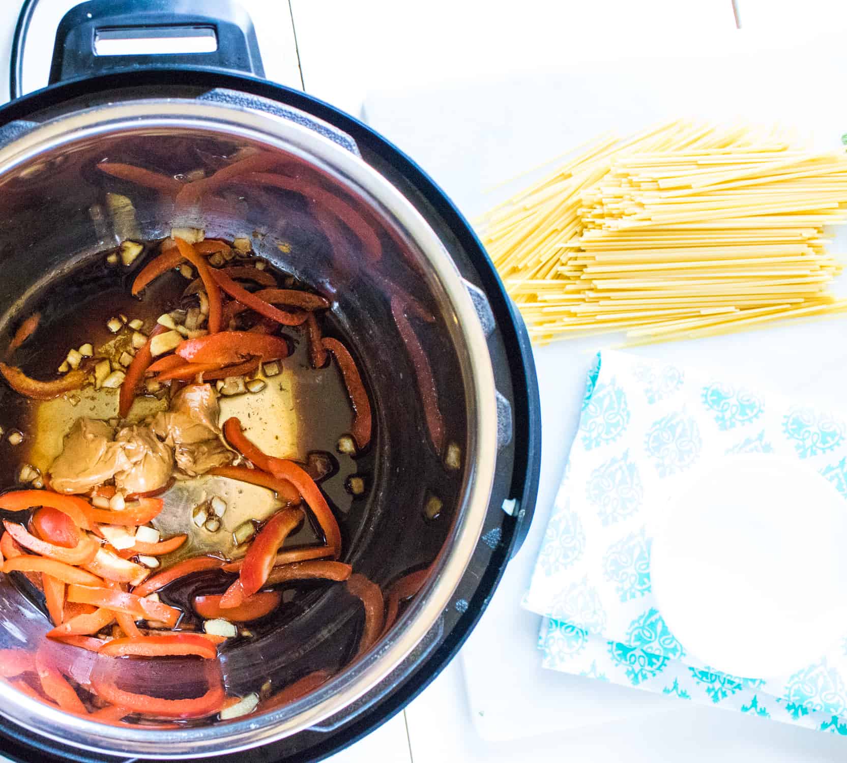 Uncooked noodles and a blue patterned napkin on a table next to an Instant Pot with ingredients for Thai Peanut Noodles inside
